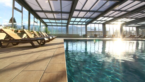 Piscina interior iluminada por el sol con sillones de descanso y vistas a montañas.
