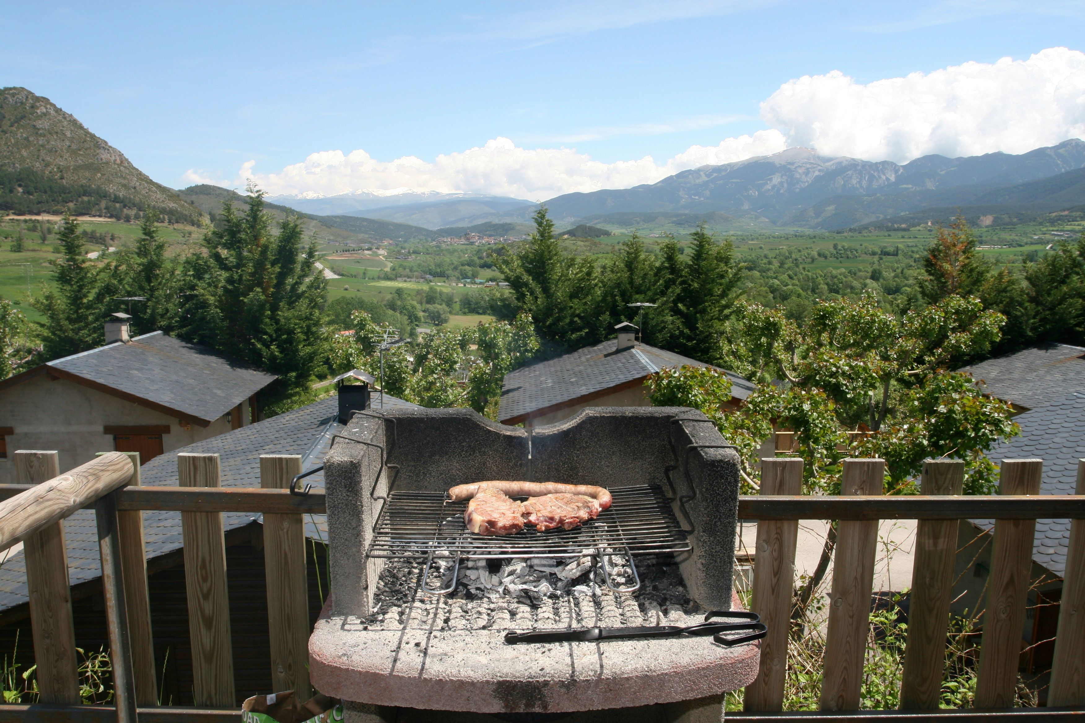 A barbecue grill with meat, surrounded by a wooden fence, set against a backdrop of mountains and a clear sky.