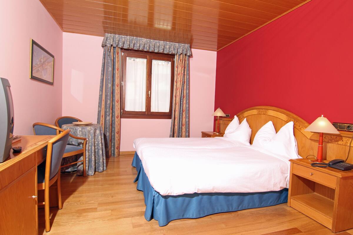 A hotel room with a bed, a desk, a chair, a table, and a nightstand with a lamp.