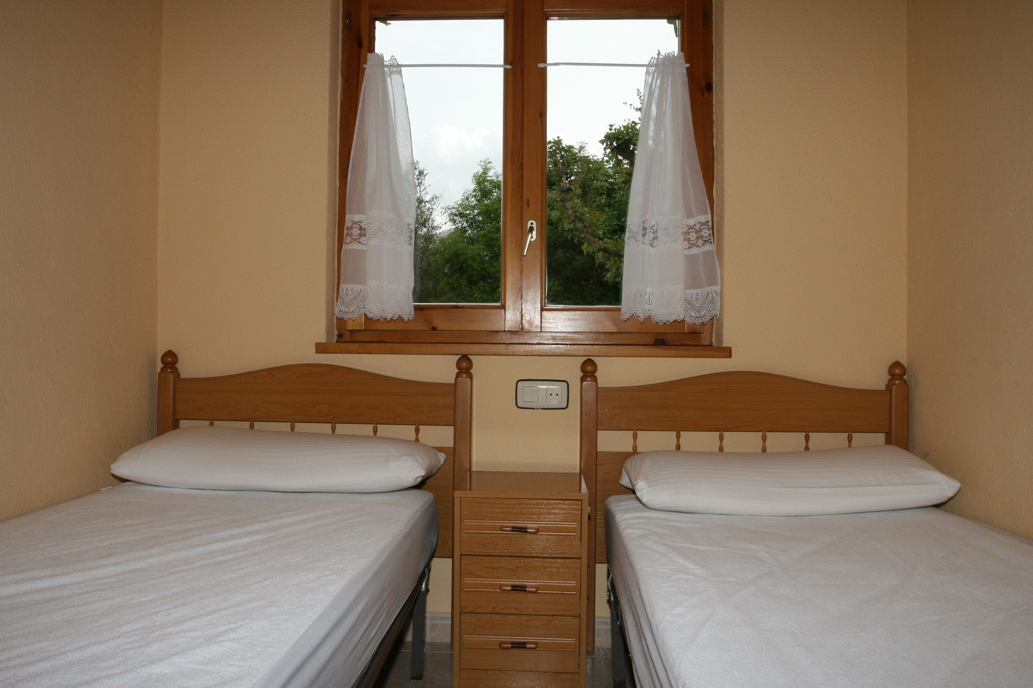 Una habitación con dos camas individuales, un cabecero de madera, una ventana con cortinas blancas y un pequeño aparador de madera entre las camas.