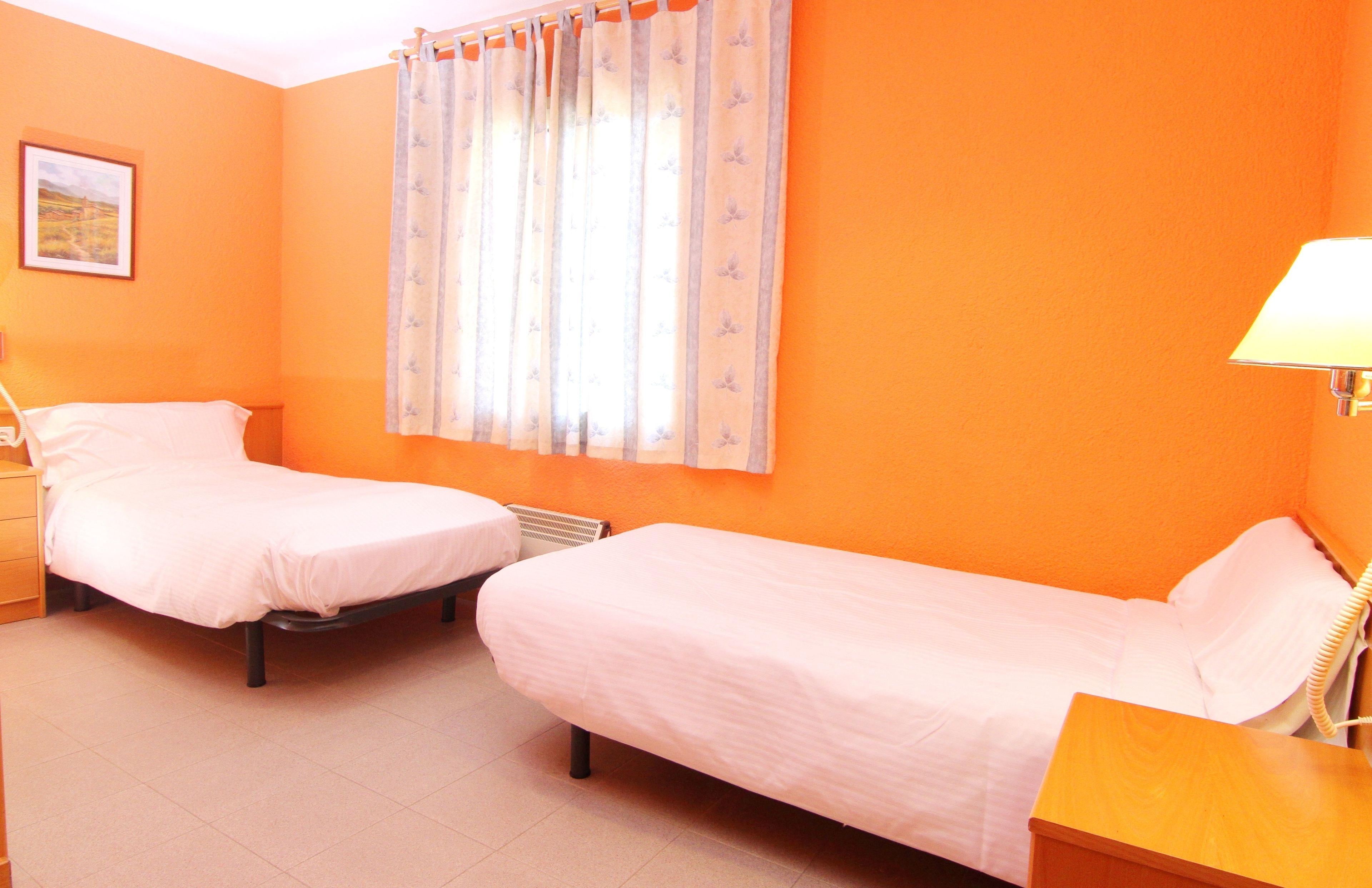 A room with two single beds, a bedside table with a lamp, a window with curtains, and a picture on the wall.