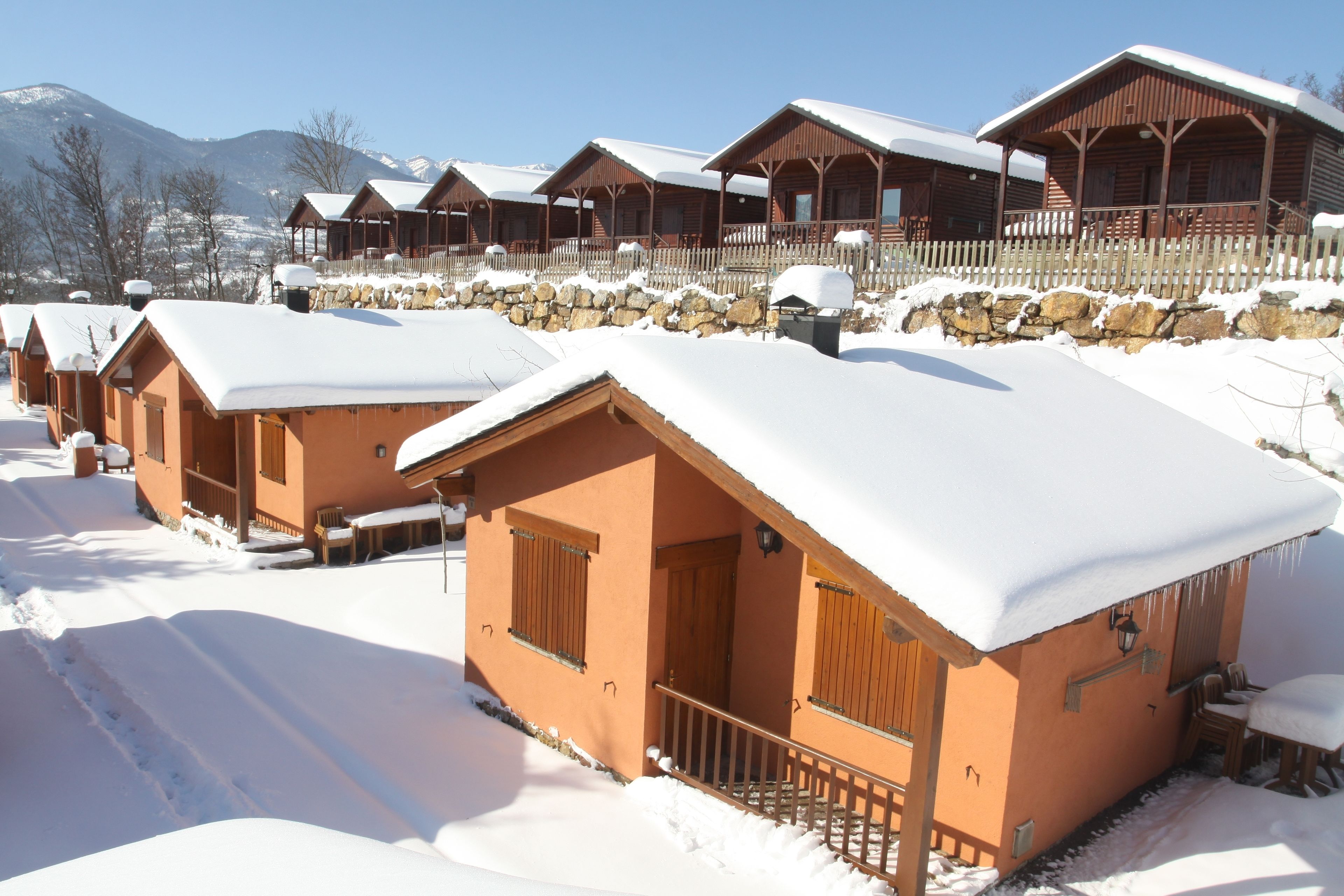 A row of wooden cabins with snow-covered roofs and a stone wall in the background.