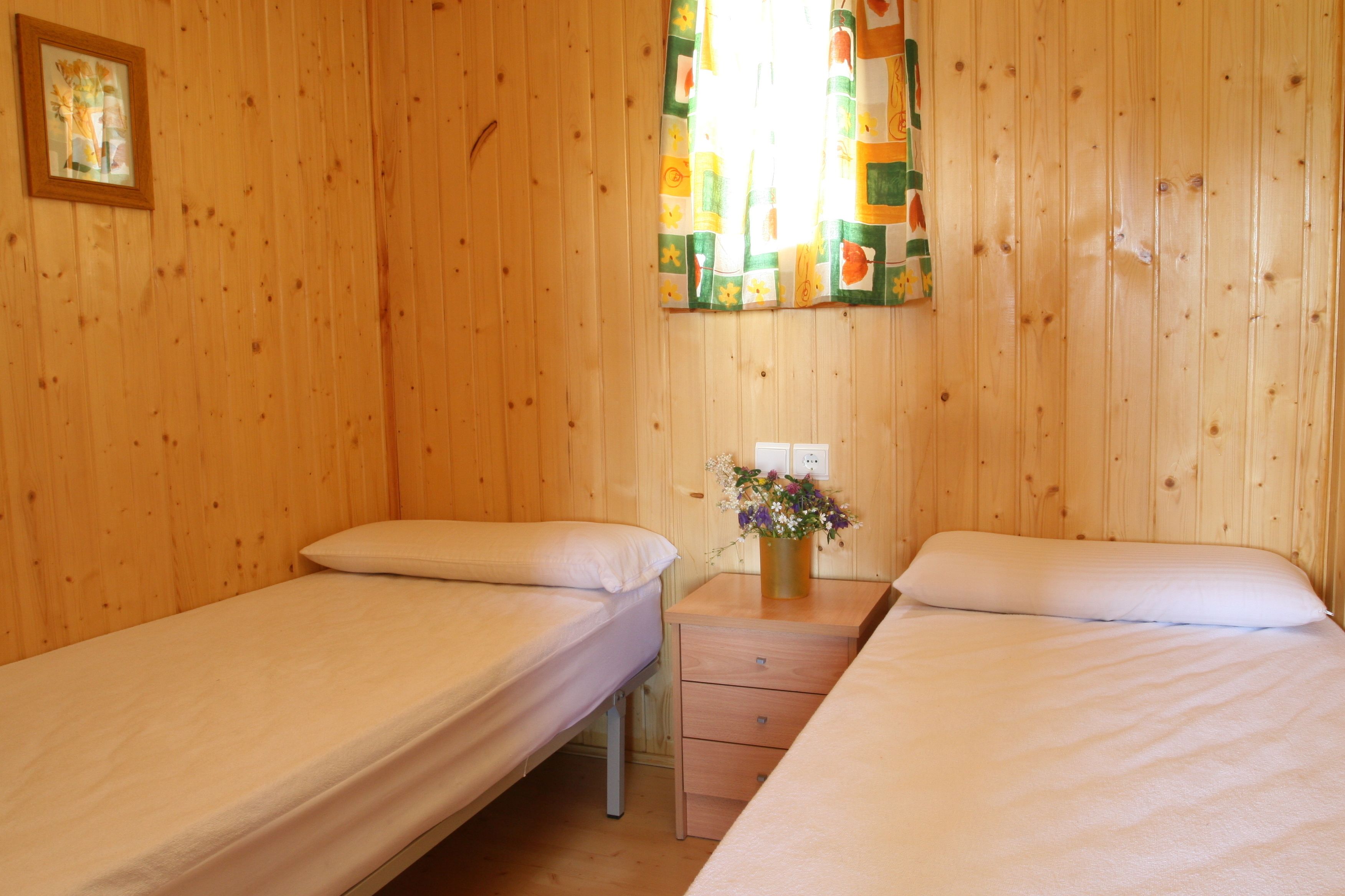 Une chambre avec deux lits simples, des murs en bois, une petite table de chevet en bois ornée d’un vase de fleurs et une fenêtre avec des rideaux.