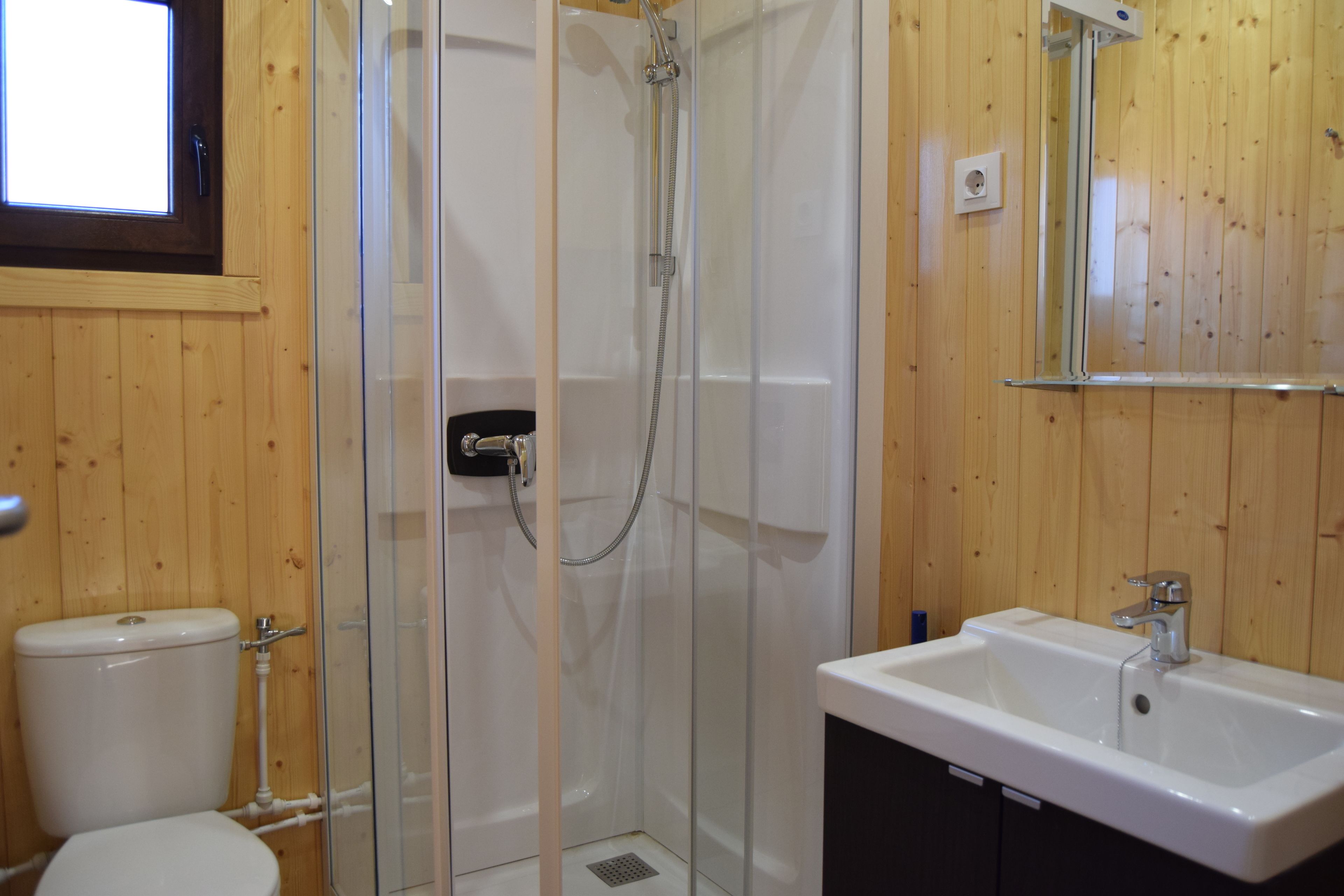 Un baño con ducha envuelta en vidrio, un lavabo blanco y una pared de madera.