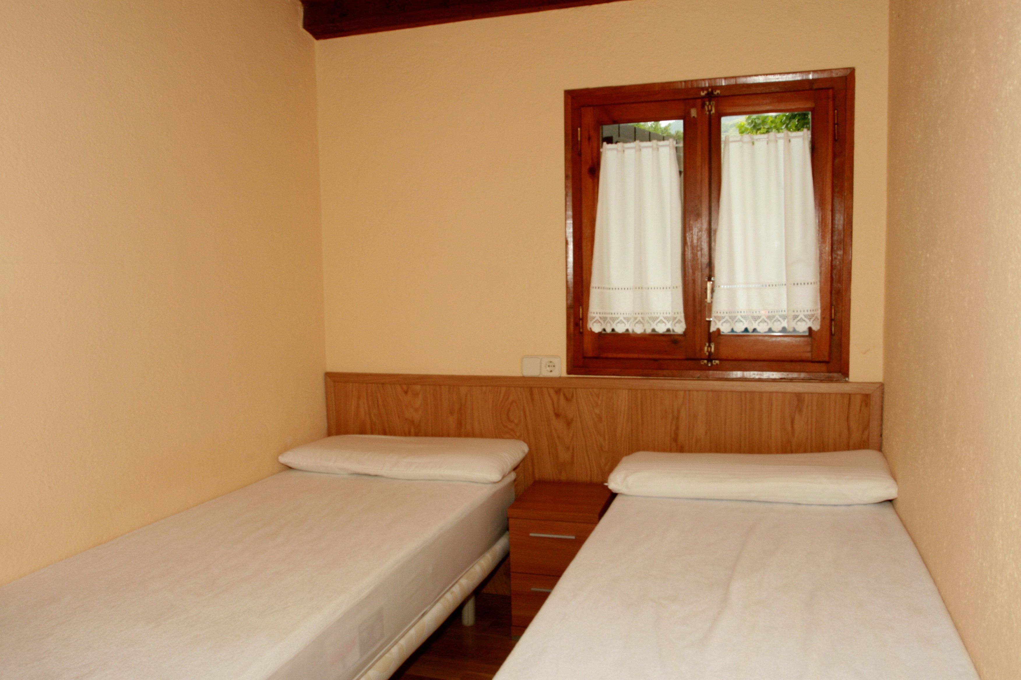 Una habitación pequeña con dos camas individuales, un banco de madera y una ventana con cortinas.