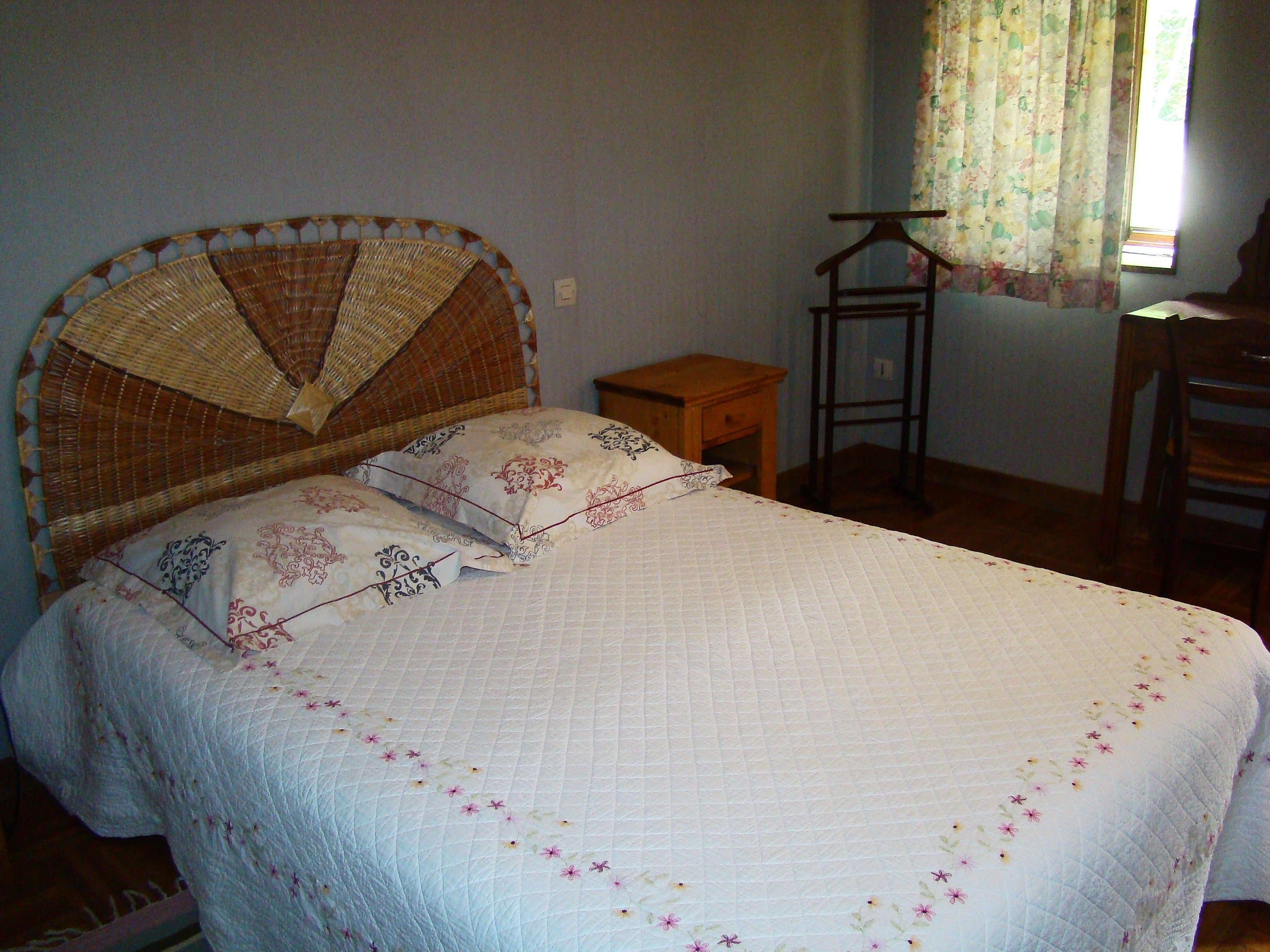 Une chambre à coucher avec un lit, une table de chevet en bois, une chaise et une fenêtre ornée de rideaux à motifs floraux.