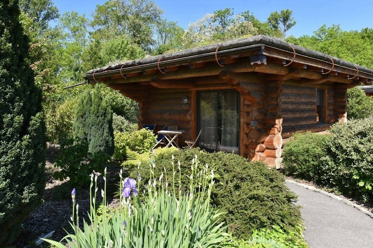 A log cabin with a thatched roof, surrounded by lush greenery and flowering plants.
