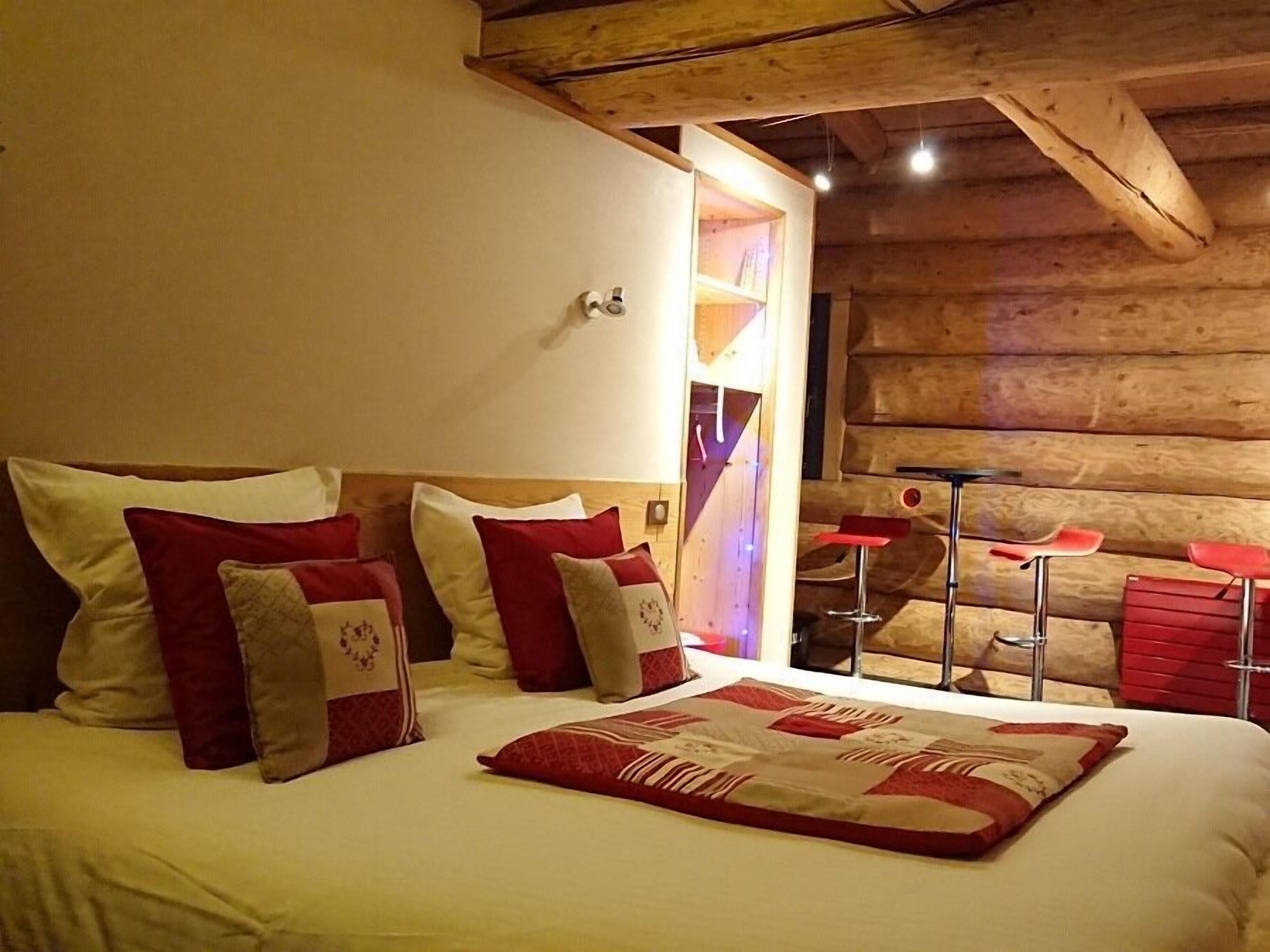 A cozy room with a wooden ceiling, a bed with red and white bedding, and a small bar area with red stools.