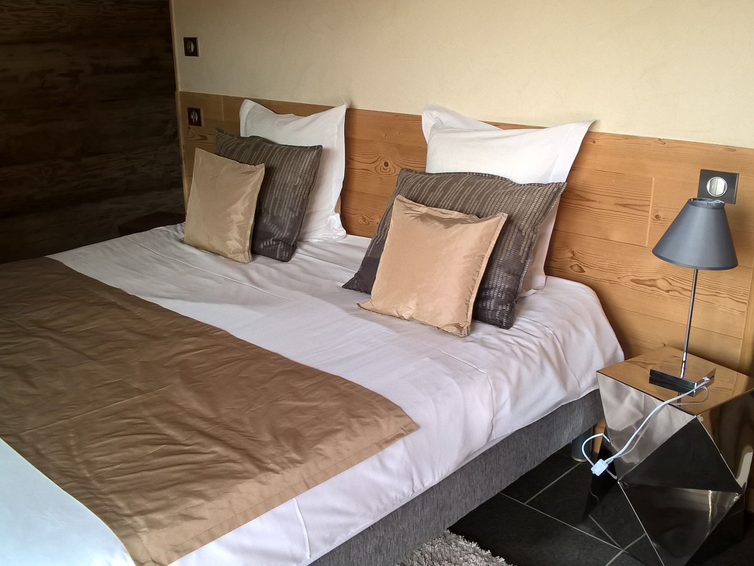 Un letto rifatto con cura, una coperta marrone, una testiera in legno e un comodino con una lampada e un cavo di ricarica.