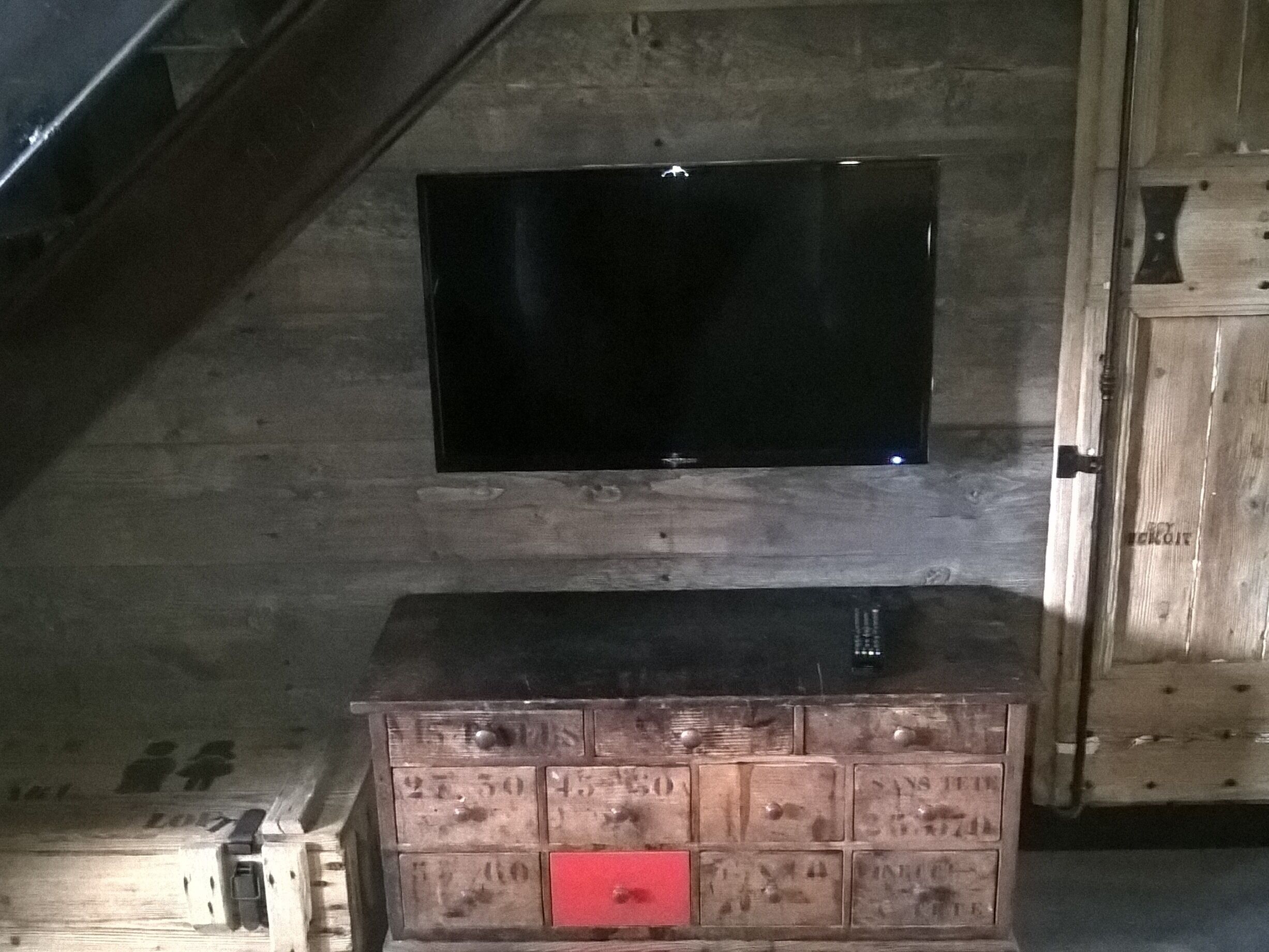 A wooden chest of drawers with a red lock, a black television mounted on the wall, and a wooden cabinet to the right.