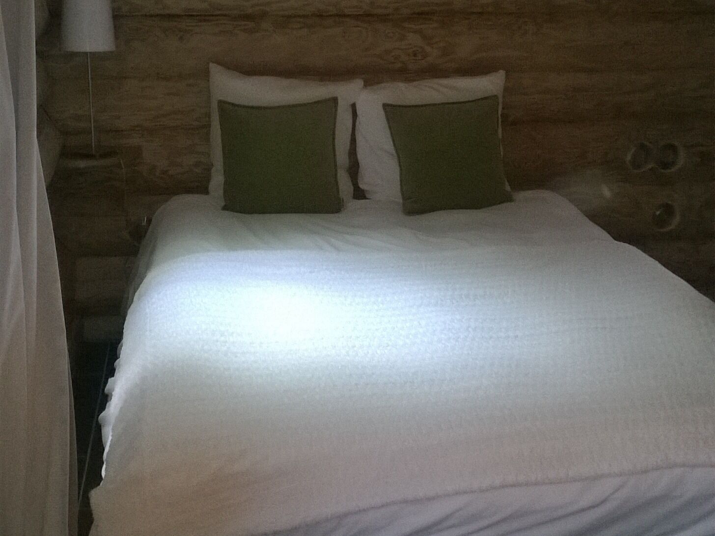 A bed with white bedding and two green pillows in a room with wooden walls.