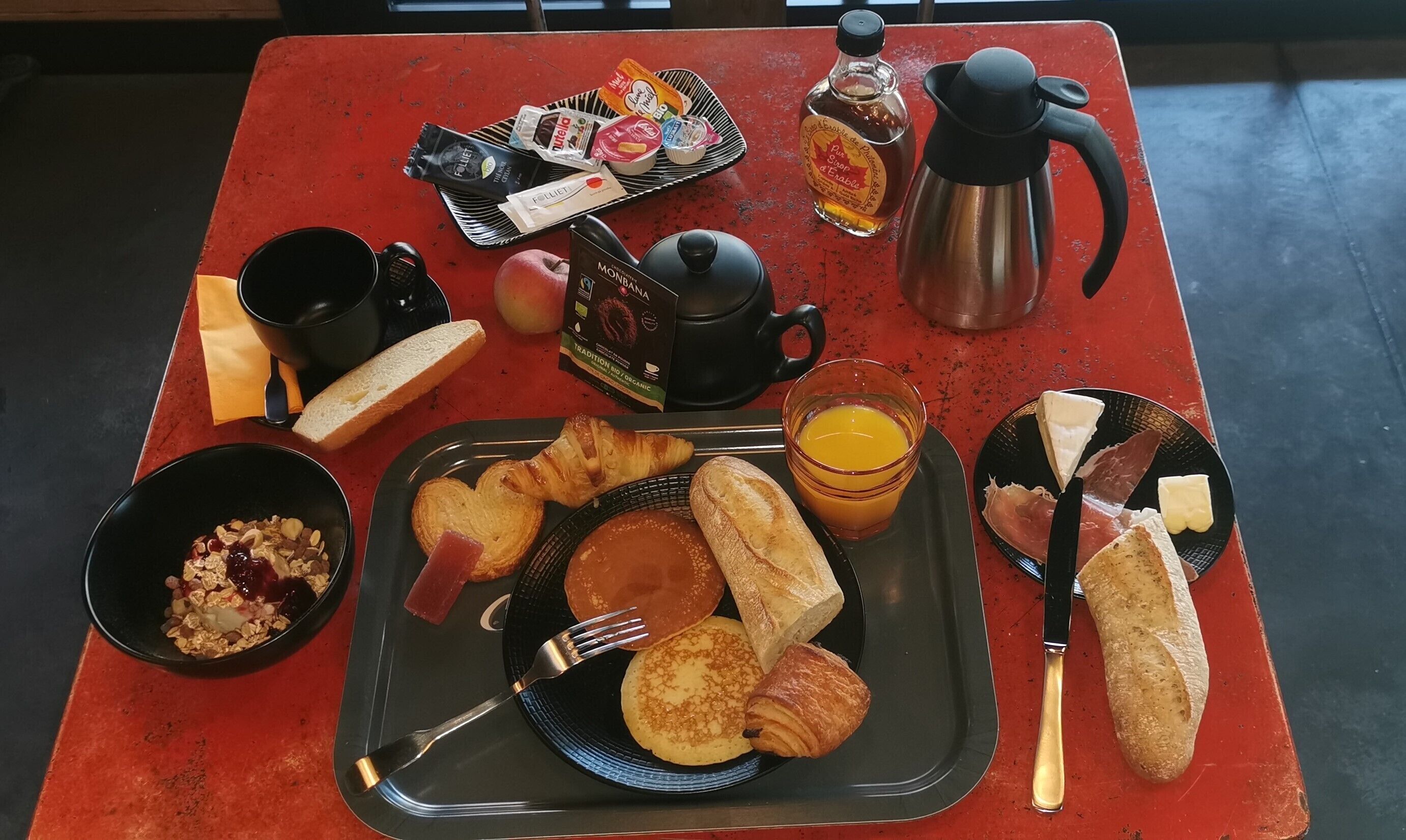 A breakfast table with various food items, a coffee pot, and a bottle of syrup.