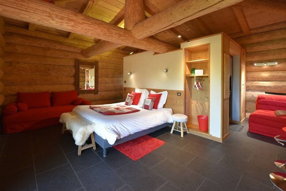 A log cabin bedroom with a bed, red sofas, and a wooden ceiling.