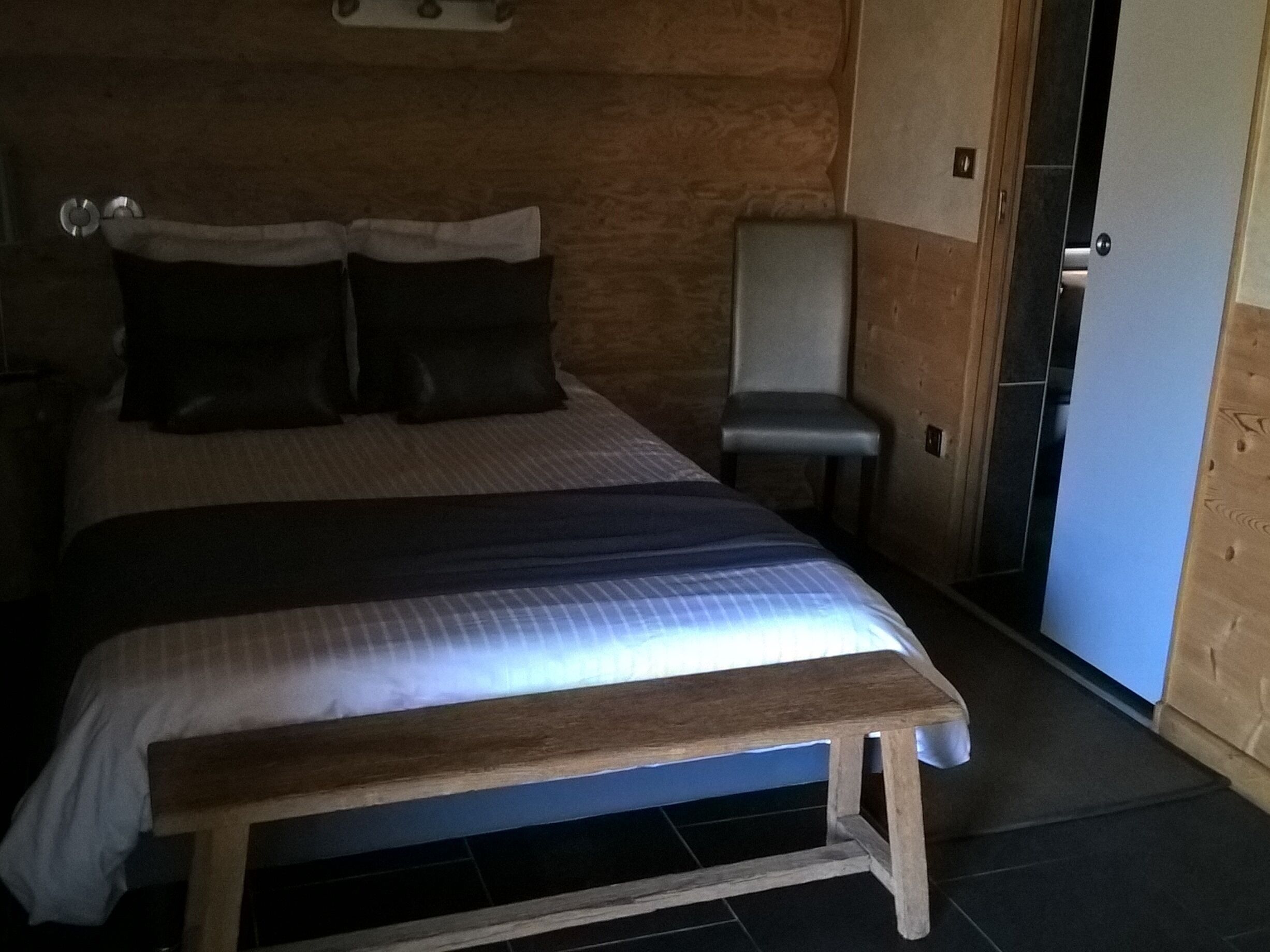 Een slaapkamer met een bed, kussens, een bankje, een stoel en een deur.