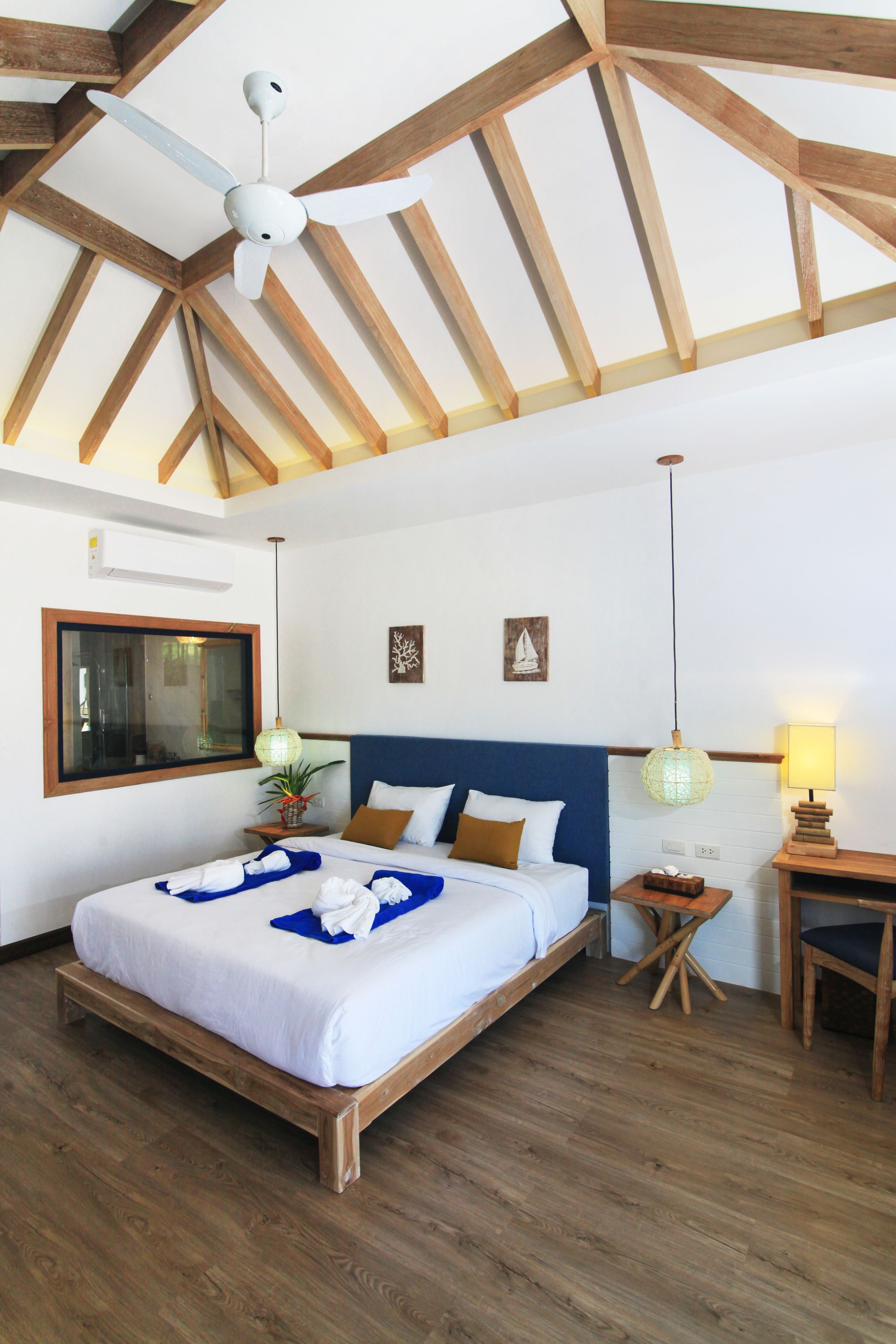 A bedroom with a wooden ceiling, a bed with white bedding, a blue headboard, a bedside table with a lamp, and a window with a view.