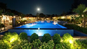 A nighttime view of a swimming pool with tiled blue flooring, surrounded by well-lit lounge areas and palm trees.