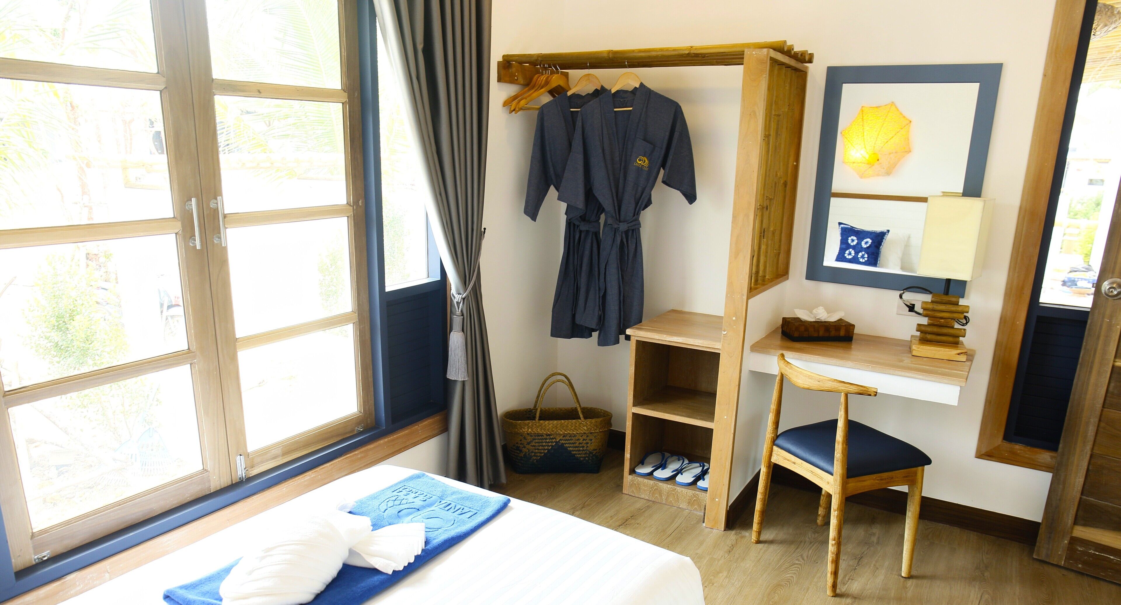 A hotel room with a bed, a wooden chair, a small table, a shelf with slippers, a basket, and a robe hanging on a rack.