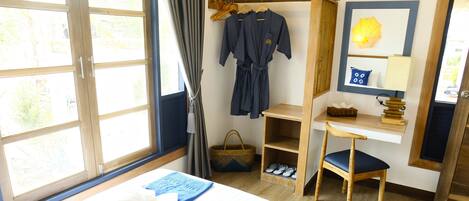 A hotel room with a bed, a wooden chair, a small table, a shelf with slippers, a basket, and a robe hanging on a rack.