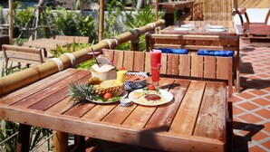 An outdoor dining area with wooden tables and chairs, surrounded by lush greenery and a clear blue sky.
