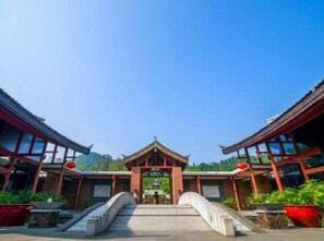 Une porte chinoise traditionnelle, avec un pont menant à celle-ci, entourée de plantes en pot et sous un ciel dégagé.
