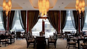 Un comedor con varias mesas y sillas, amplias ventanas con cortinas, y una lámpara de araña moderna.