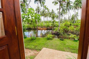 A view through a wooden door of a lush garden with a pond, surrounded by various tropical plants and palm trees. - Yoho Milkyway Holiday Resort (Unawatuna)