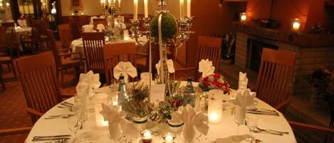 Une salle à manger élégante, avec des tables rondes dressées pour le repas, des bougies et une cheminée en arrière-plan.