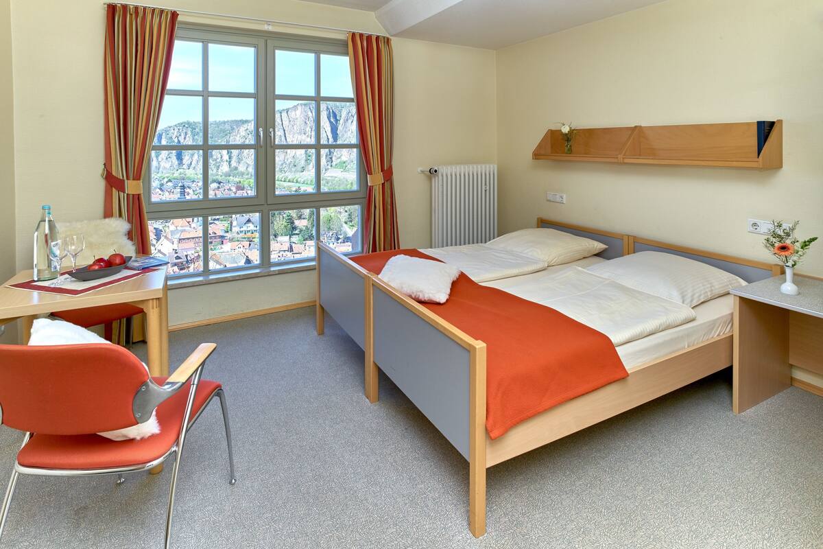 Habitación de hotel con cama, escritorio con silla, ventana con cortinas, radiador y una estantería pequeña.