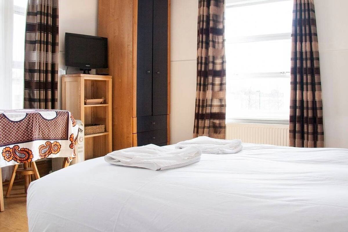 A hotel room with a large bed, a TV on a wooden stand, a wardrobe, and a window with curtains.