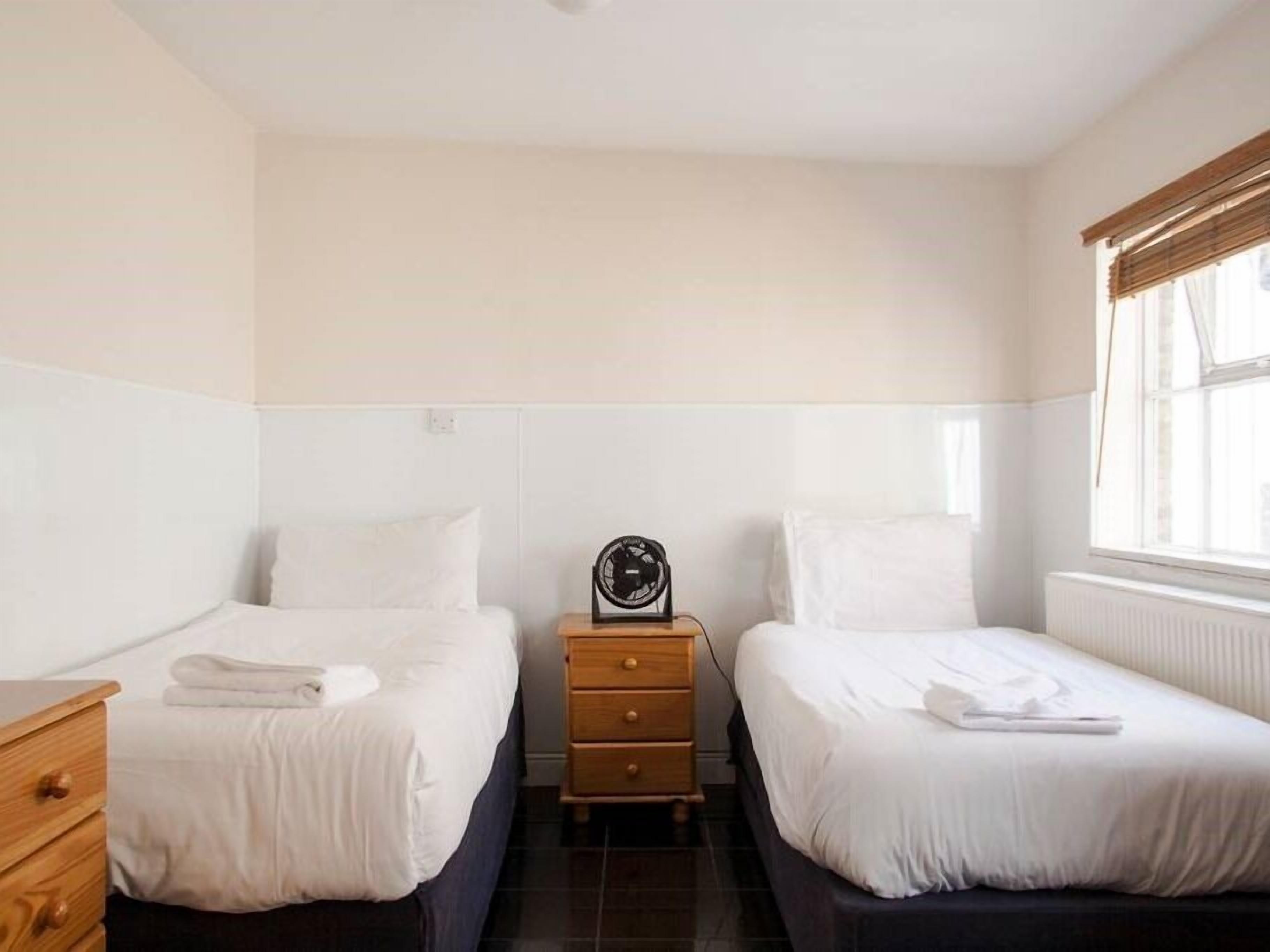 Une chambre avec deux lits simples, une commode en bois, une table de chevet avec une horloge et une fenêtre avec des stores.