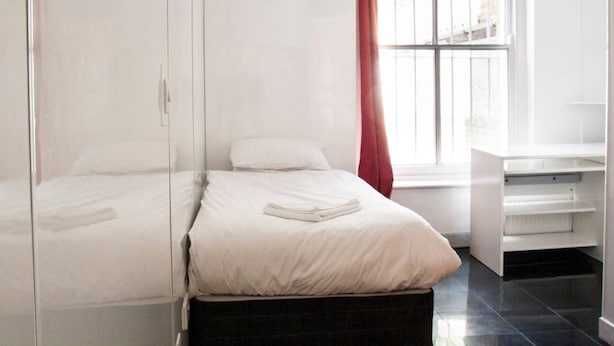 A bedroom with two beds, a window with red curtains, and a white wardrobe. - Caledonian Studio Flats (London)