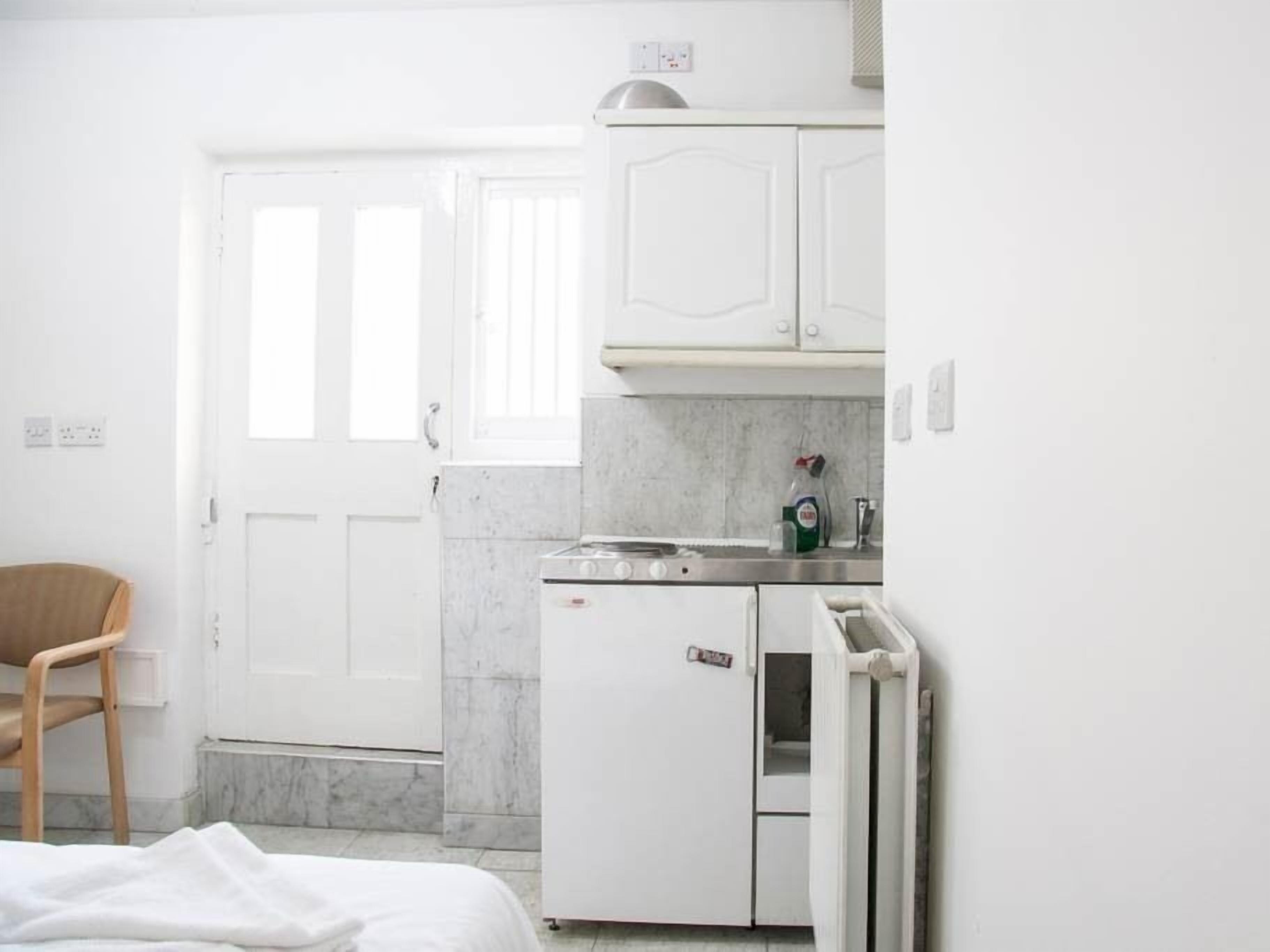 Une petite pièce blanche, avec un lit, une chaise en bois, un coin cuisine équipé d’un réfrigérateur, d’une cuisinière et de placards, et une porte avec une fenêtre.
