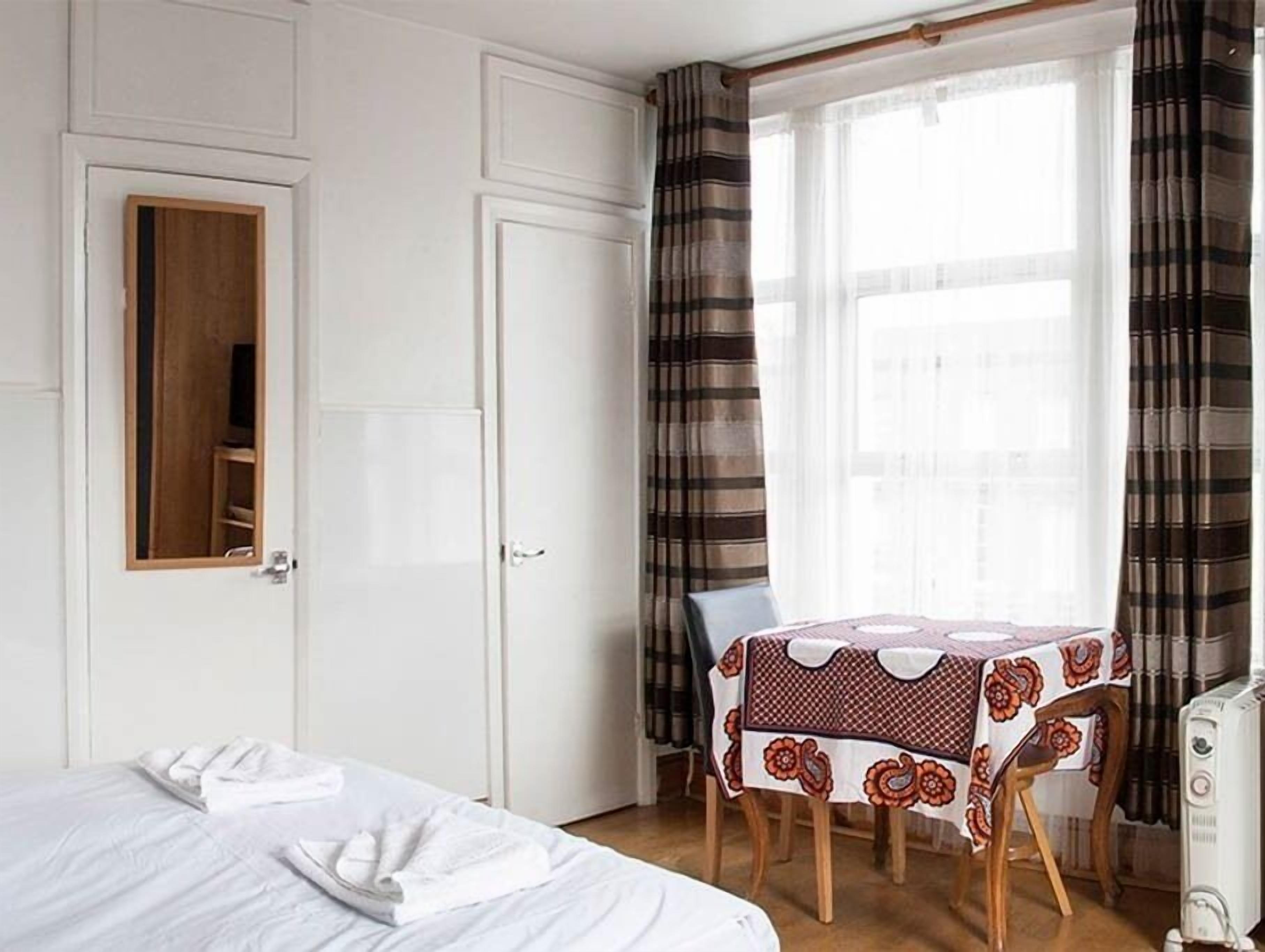 A hotel room with a bed, a table with a patterned tablecloth, a chair, a radiator, and a window with curtains.