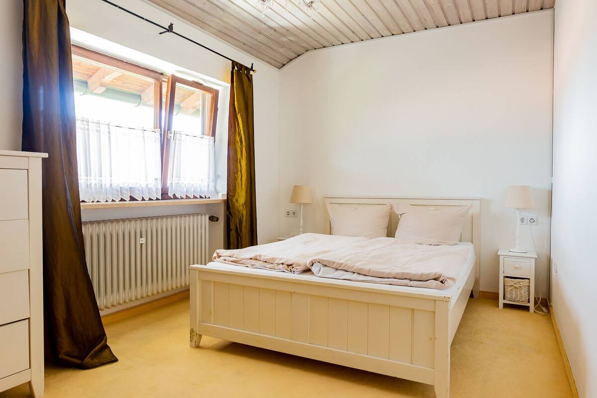A bedroom with a bed, bedside lamps, a window with curtains, a radiator, and a cabinet.
