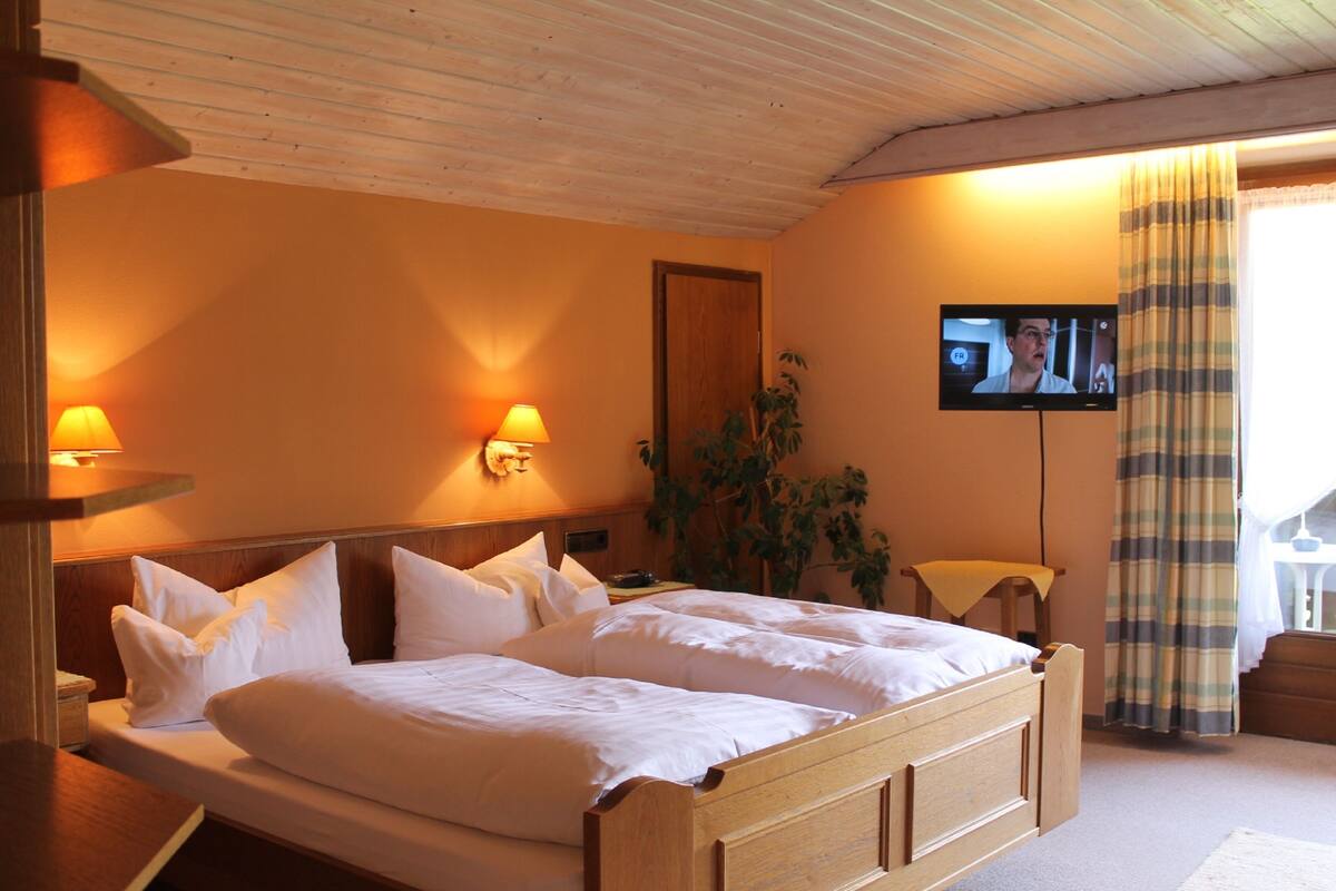 A double bed with white linens, a wooden headboard, two wall-mounted lamps, a television on the wall, and a window with curtains.
