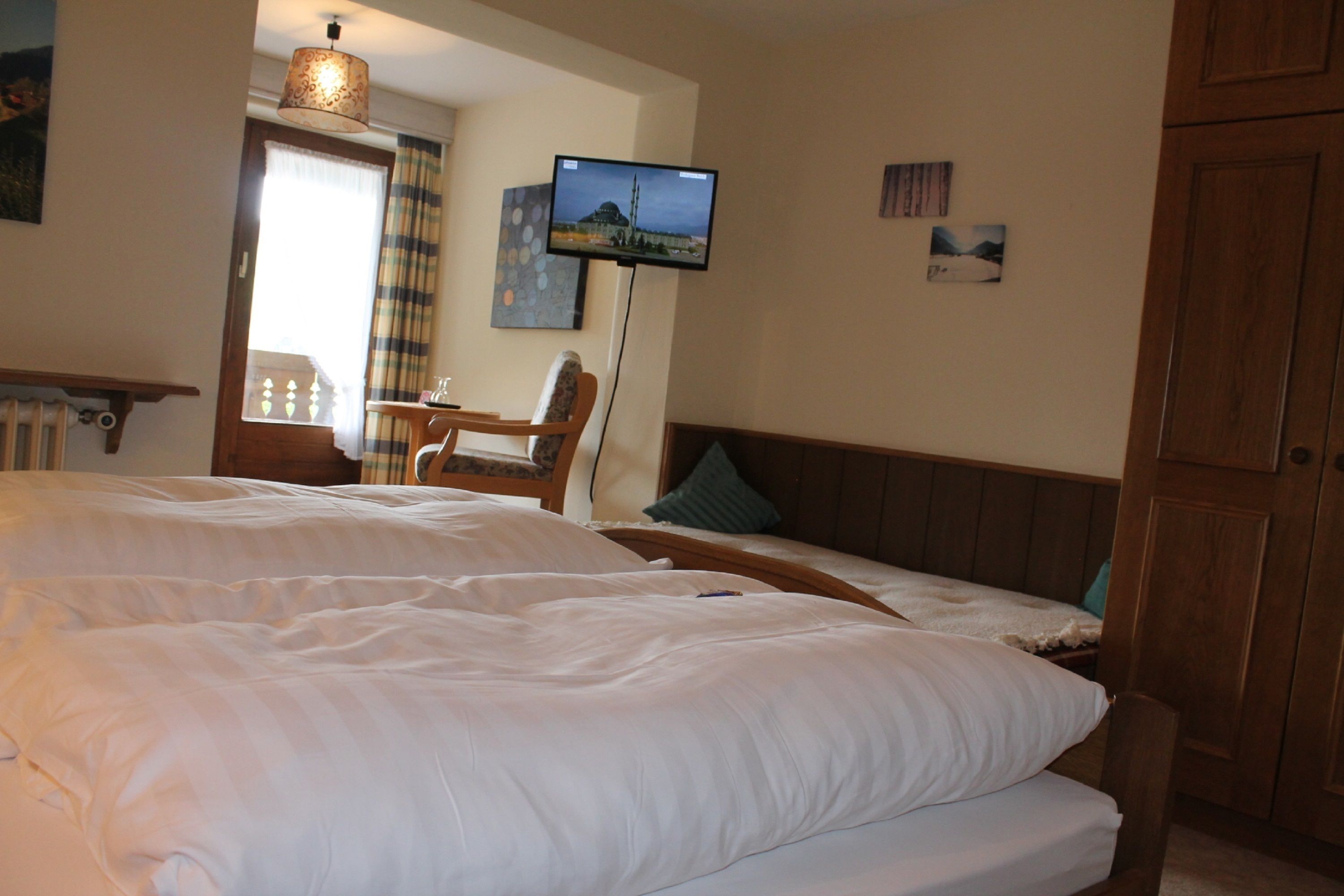 A hotel room with a large bed, a television mounted on the wall, a wooden headboard, and a window with curtains.