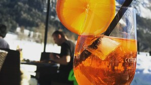 Una mano sosteniendo un vaso con una bebida, hielo y una rodaja de naranja, con una montaña nevada y una lámpara al fondo.