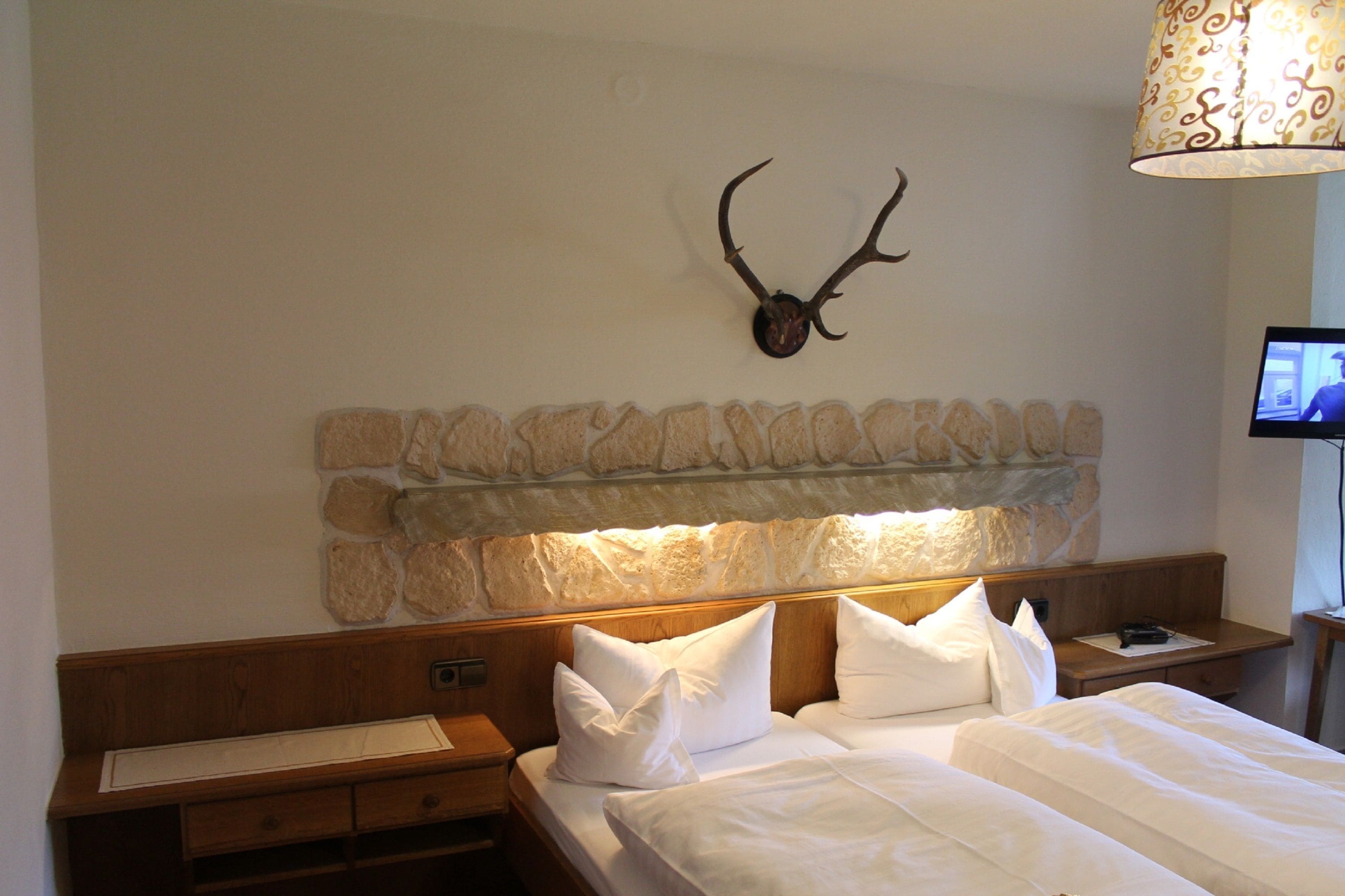 Habitación de hotel con cabecera de piedra, cabeza de venado montada en la pared, cama con ropa blanca y un televisor sobre una mesita de noche de madera.