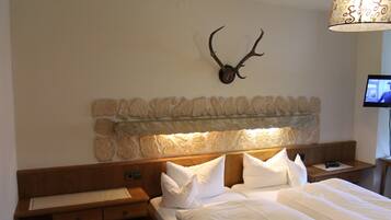 Habitación de hotel con cabecera de piedra, cabeza de venado montada en la pared, cama con ropa blanca y un televisor sobre una mesita de noche de madera.