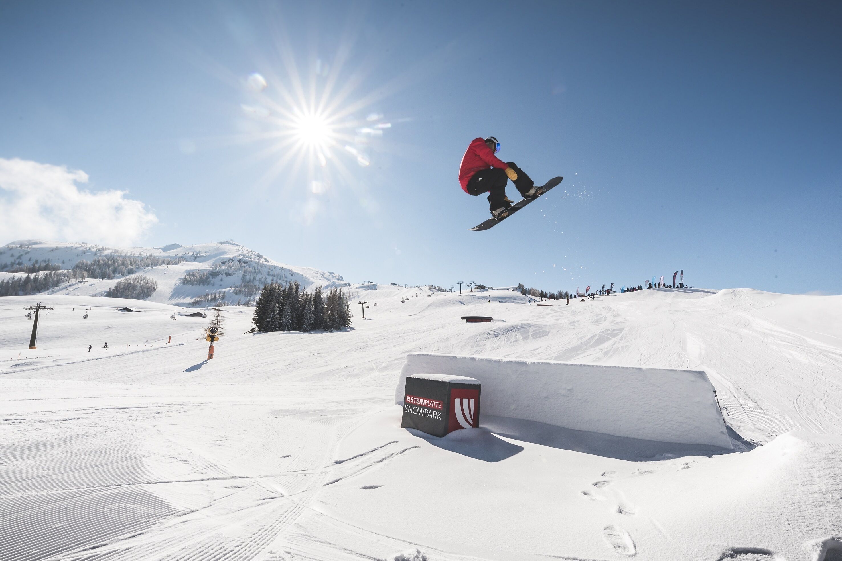 Un snowboardeur effectuant une figure acrobatique dans un parc à neige, avec un ciel dégagé et un paysage enneigé.