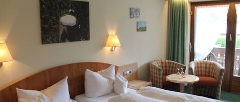 A hotel room with two beds, a small table with a chair, and a view of the outdoors.