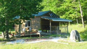 Une cabane en rondins dotée d’un porche, entourée d’arbres et d’une zone herbeuse.