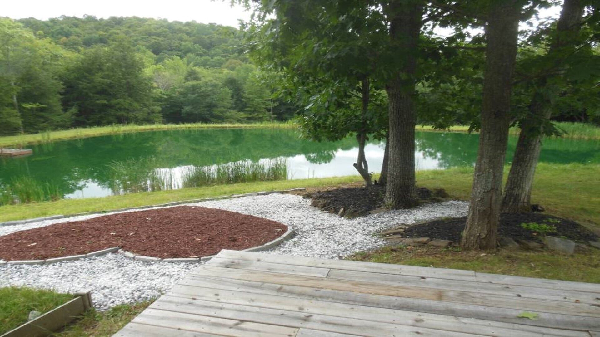 Eine Holzterrasse führt zu einem Teich, der von Bäumen und einer Rasenfläche umgeben ist.