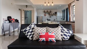 Uma sala de estar moderna com um sofá preto, almofadas estampadas e uma almofada com a bandeira da Union Jack. Uma mesa de jantar com toalha de mesa é visível ao fundo.