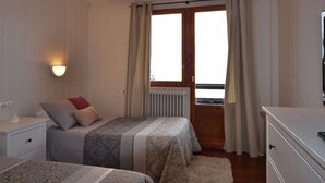Une chambre avec deux lits, une table de chevet, une commode, un radiateur et une fenêtre avec des rideaux.
