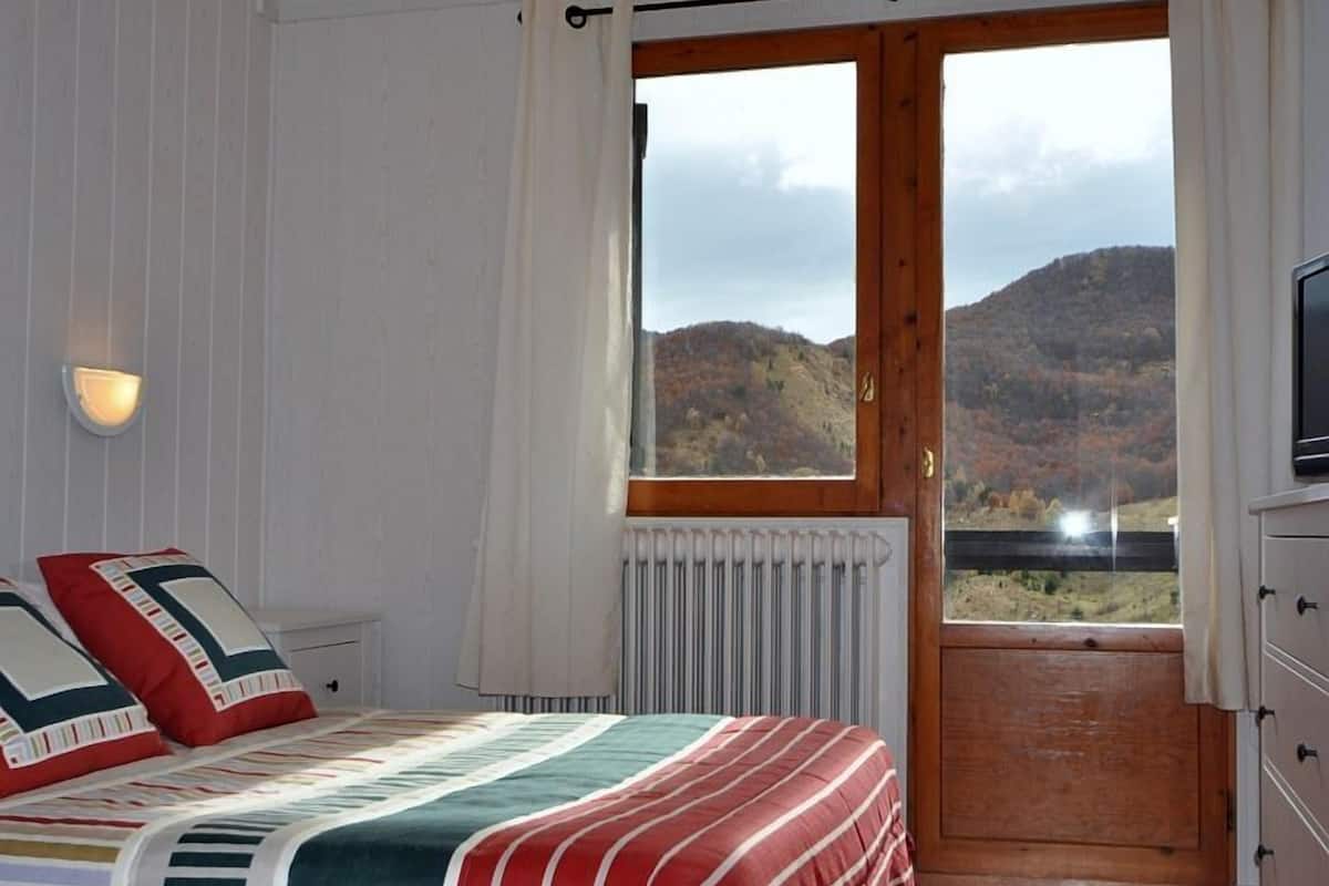 Une chambre à coucher comprenant un lit, une table de chevet, un téléviseur, une fenêtre donnant sur les montagnes et une porte en bois.