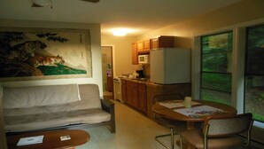 A compact living space with a sofa, a dining table with chairs, a kitchen area with a microwave and stove, and a large window with a view of greenery.