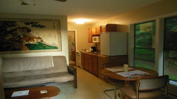 A compact living space with a sofa, a dining table with chairs, a kitchen area with a microwave and stove, and a large window with a view of greenery.