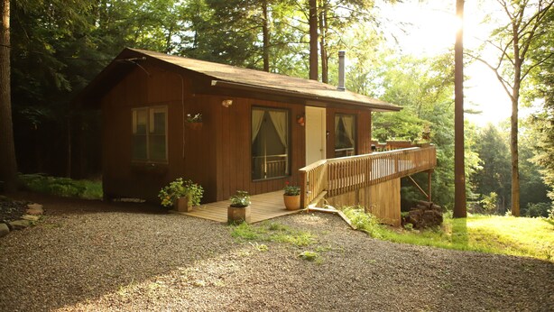A small wooden cabin with a deck, surrounded by trees and a gravel driveway.