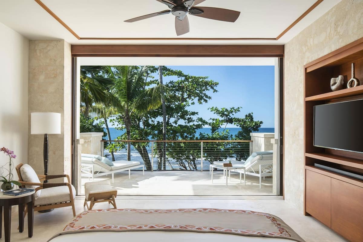 Una habitación moderna con un gran ventanal corredero que da a un balcón con vistas al océano y palmeras.